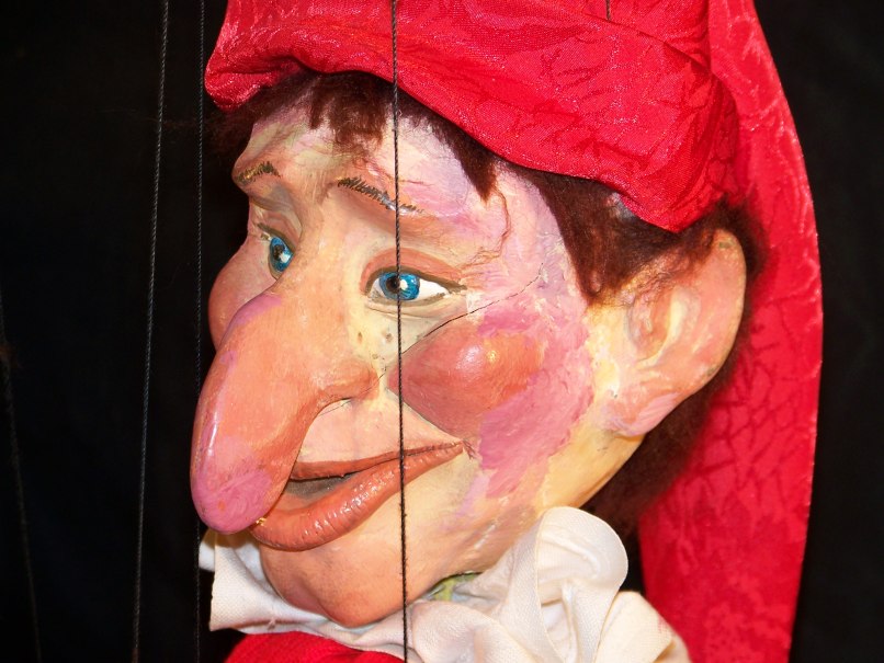 Nahaufnahme einer Kasperle-Puppe mit roter Mütze, großen blauen Augen und markanter Nase. Der Hintergrund ist dunkel., © Nellys Puppentheater