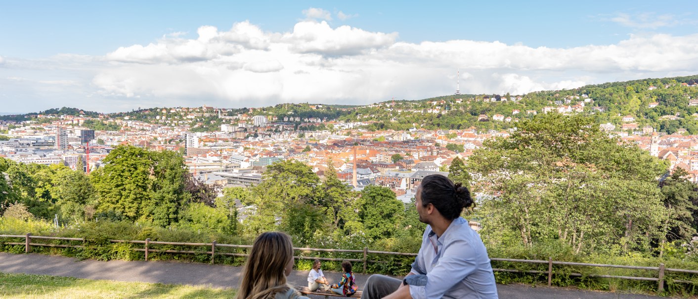 Zwei Personen sitzen auf einer Wiese und blicken auf die Stadt Stuttgart. Im Hintergrund sind Hügel und ein blauer Himmel mit Wolken zu sehen., © © Stuttgart-Marketing GmbH, Martina Denker Zwei Personen sitzen auf einer Wiese und blicken auf die Stadt Stuttgart. Im Hintergrund sind Hügel und ein blauer Himmel mit Wolken zu sehen., © © Stuttgart-Marketing GmbH, Martina Denker