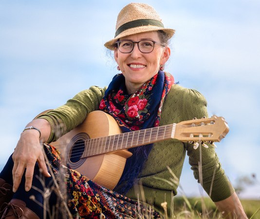Eine Person mit Strohhut und Brille sitzt lächelnd auf einer Wiese, hält eine Gitarre und trägt bunte Kleidung., © Rosenau Kultur e.V. Eine Person mit Strohhut und Brille sitzt lächelnd auf einer Wiese, hält eine Gitarre und trägt bunte Kleidung., © Rosenau Kultur e.V.