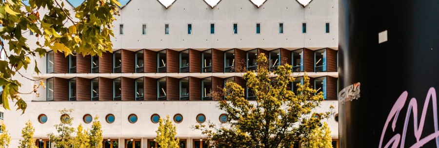 Moderne Bibliothek mit markanter geometrischer Fassade, umgeben von B&auml;umen. Ein schwarzer Pfeiler mit Graffiti ist rechts im Bild sichtbar., &copy; Stuttgart Marketing GmbH, Sarah Schmid