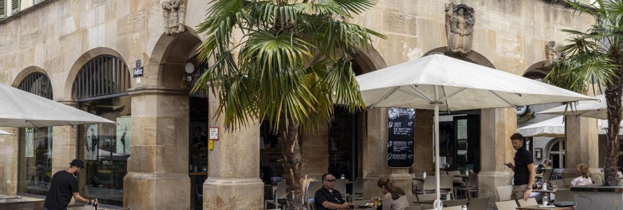 Café mit Tischen und Stühlen unter Sonnenschirmen vor einem historischen Gebäude mit Arkaden. Palmen und Menschen im Außenbereich., © SMG, Sarah Schmid