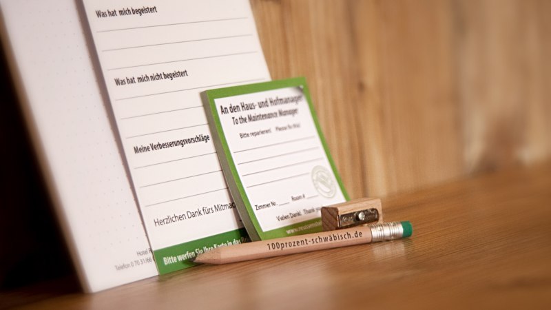 A pencil labeled "100prozent-schwäbisch.de", a pencil sharpener and forms for opinions and repair requests on a wooden table., © TOMAS A pencil labeled "100prozent-schwäbisch.de", a pencil sharpener and forms for opinions and repair requests on a wooden table., © TOMAS