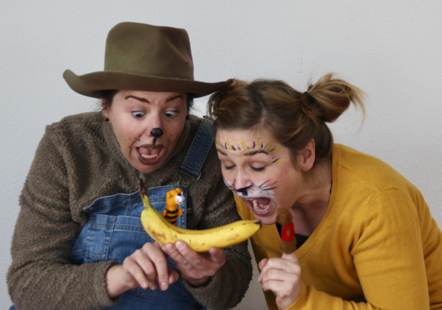 Zwei verkleidete Personen mit Tiergesichtsbemalung bestaunen eine Banane, auf der ein kleines Tiger-Spielzeug sitzt. Sie wirken überrascht und erfreut., © Theater Kopfkonfetti