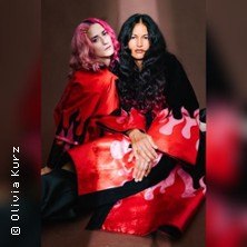 Zwei Personen mit langen Haaren und auff&auml;lliger Kleidung mit Flammenmuster posieren vor einem neutralen Hintergrund., &copy; links im Bild