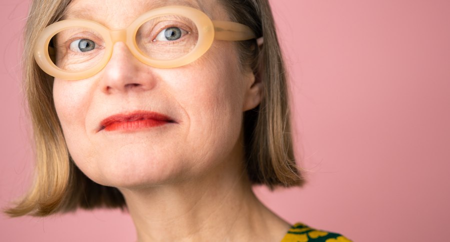 Eine Frau mit blonden Haaren und auff&auml;lliger, runder Brille blickt in die Kamera. Der Hintergrund ist rosa, ihr Oberteil hat ein gelbes Muster., &copy; Foto: Annette Koroll