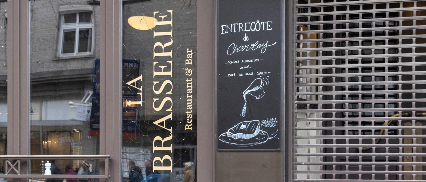 Eingang von 'La Brasserie' mit Men&uuml;tafel, die Entrec&ocirc;te de Charolais mit Pommes und Caf&eacute; de Paris Sauce anbietet., &copy; SMG, Sarah Schmid
