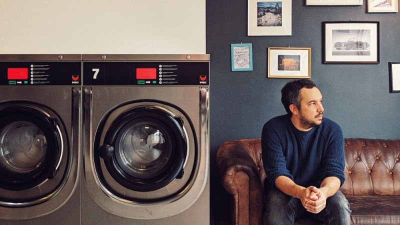 Ein Mann sitzt auf einem Ledersofa in einem Waschsalon neben Waschmaschinen. An der Wand hängen Bilder und eine Uhr., © Veranstalter Ein Mann sitzt auf einem Ledersofa in einem Waschsalon neben Waschmaschinen. An der Wand hängen Bilder und eine Uhr., © Veranstalter