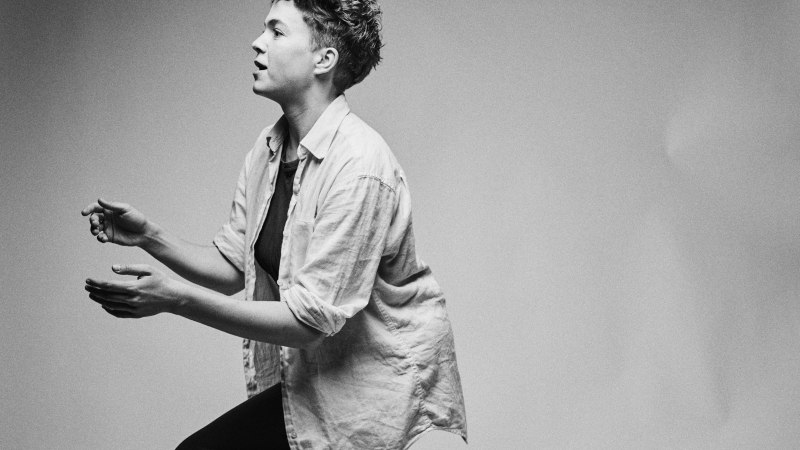 Schwarz-weiß Bild einer Person, die seitlich sitzt und gestikuliert. Die Person trägt ein Hemd und hat lockiges Haar., © Felix Groteloh Schwarz-weiß Bild einer Person, die seitlich sitzt und gestikuliert. Die Person trägt ein Hemd und hat lockiges Haar., © Felix Groteloh