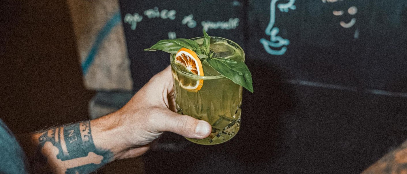 Eine t&auml;towierte Hand h&auml;lt ein gr&uuml;nes Getr&auml;nk mit Basilikum und Orangenscheibe. Im Hintergrund ist eine Tafel mit Zeichnungen und Schrift zu sehen., &copy; Bruno, Stuttgart