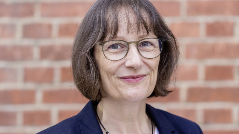 Eine Frau mit Brille und braunen Haaren lächelt vor einer Ziegelwand. Sie trägt einen dunklen Blazer und eine Halskette., © arge lola