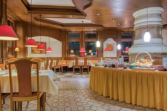 Gemütlicher Frühstücksraum mit gedeckten Tischen, roten Lampen und einem Buffet mit Gebäck. Holzwände und ein großer Ofen schaffen eine warme Atmosphäre., © Centro Hotels