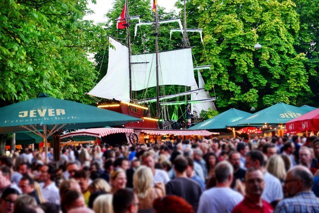 Belebter Fischmarkt mit Menschenmenge, Segelschiff im Hintergrund und grünen Sonnenschirmen. Lebhafte Atmosphäre unter Bäumen., © WAGS Hamburg Events GmbH Belebter Fischmarkt mit Menschenmenge, Segelschiff im Hintergrund und grünen Sonnenschirmen. Lebhafte Atmosphäre unter Bäumen., © WAGS Hamburg Events GmbH