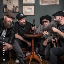 Vier Männer sitzen um einen kleinen Tisch, trinken und unterhalten sich in einem gemütlichen Raum mit Bildern an der Wand., © links im Bild