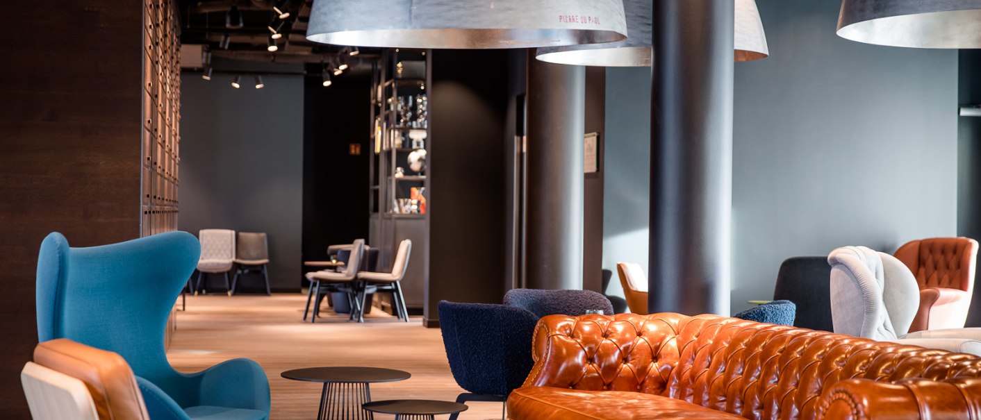 Moderne Lounge mit braunem Ledersofa, blauen Sesseln und großen, hängenden Lampen. Im Hintergrund sind weitere Sitzgelegenheiten sichtbar., © TOMAS