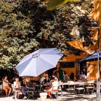 Menschen genießen das sonnige Wetter in einem Café im Freien, umgeben von Bäumen und Sonnenschirmen. Eine entspannte Atmosphäre herrscht vor., © Stuttgart-Marketing GmbH, Sarah Schmid