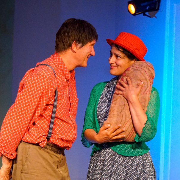 Zwei Personen in bunten Kost&uuml;men auf einer B&uuml;hne. Eine Person h&auml;lt ein B&uuml;ndel, beide l&auml;cheln sich an. Die Szene wirkt fr&ouml;hlich und lebendig., &copy; Katja Ritter