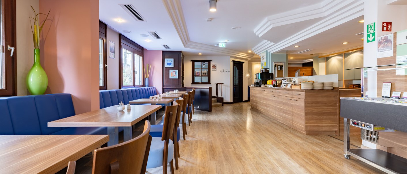A bright breakfast room with wooden tables, blue benches, and a buffet area. Large windows let in plenty of light., &copy; Novum  Hospitality
