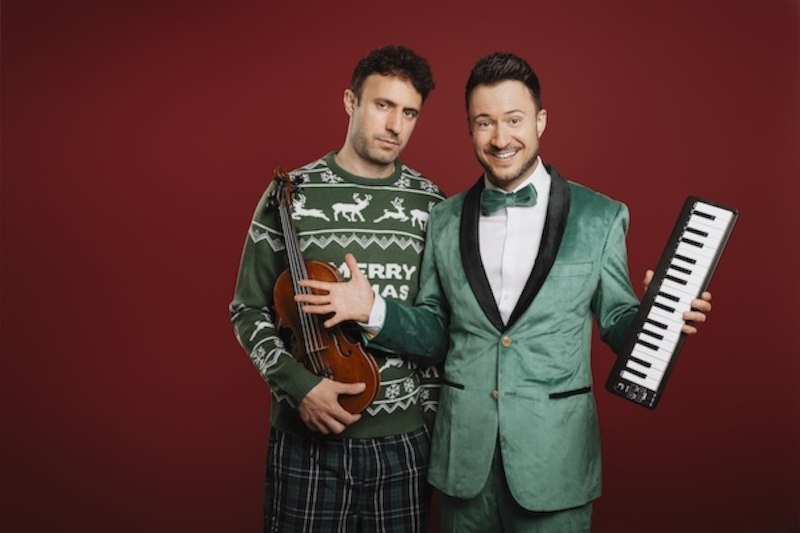 Two men pose in front of a red background. One is wearing a Christmas sweater and holding a violin, the other is wearing a green suit and holding a keyboard., &copy; Renitenztheater Stuttgart e.V.