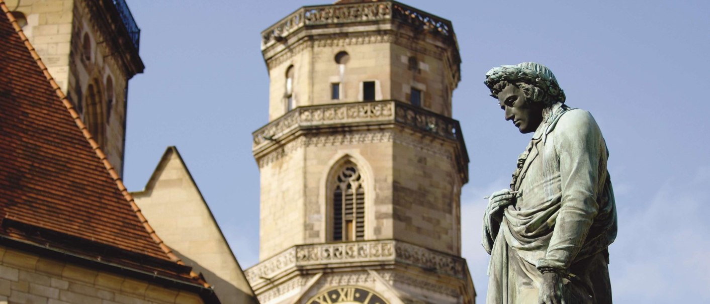 Das Schillerdenkmal steht vor einem historischen Turm mit Uhr und gotischen Fenstern, unter einem klaren blauen Himmel., © Stuttgart-Marketing GmbH
