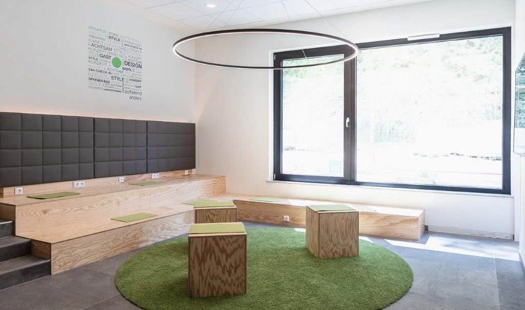 A modern lobby with wooden benches, green seat cushions, and a large window. A round rug and a circular lamp complete the design., &copy; a2 HOTELS am Quadrium