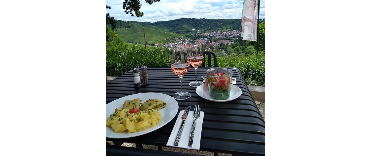 A table with two plates of food and two glasses of rosé wine. Vineyards and a village in the background., © 1819 Bistro am Wirtemberg A table with two plates of food and two glasses of rosé wine. Vineyards and a village in the background., © 1819 Bistro am Wirtemberg