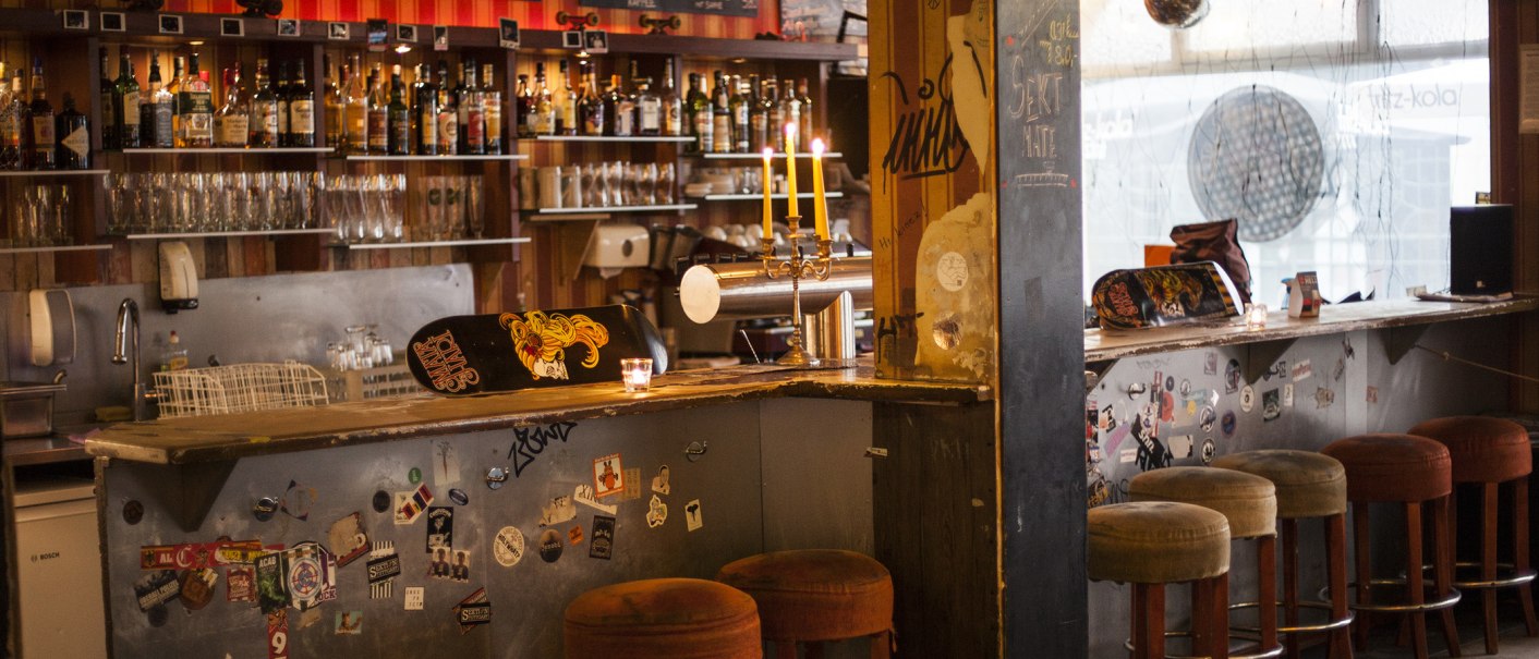 A cozy bar with skateboard decor on the counter, candlesticks and bar stools. Bottles and glasses can be seen in the background., © Mata Hari, Stuttgart
