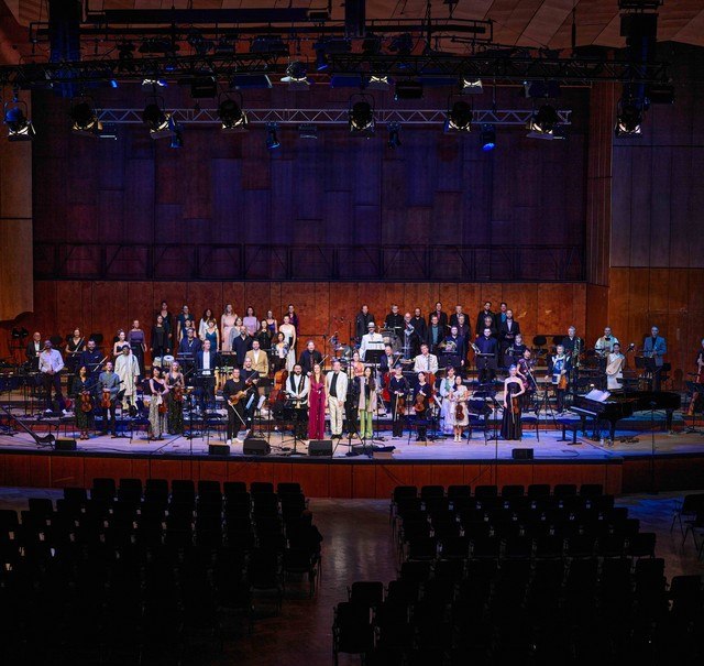 Ein Orchester mit Chor steht auf einer Bühne in einem Konzertsaal. Die Musiker tragen festliche Kleidung, der Saal ist mit leeren Stuhlreihen gefüllt., © Marc Schäfer