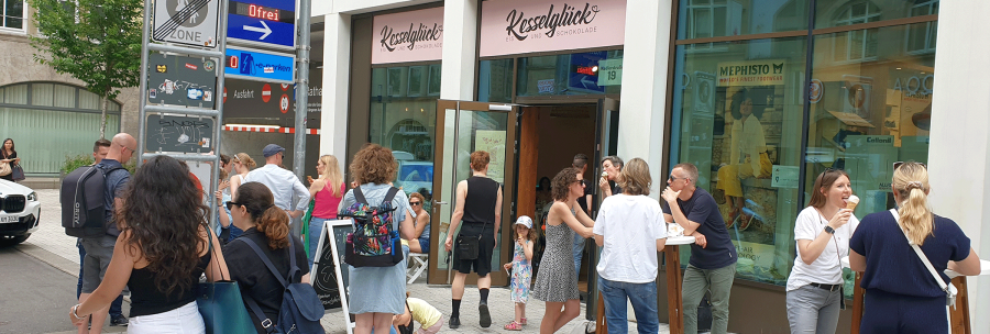 Menschen genießen Eis vor dem Eiscafé Kesselglück in einer belebten Stadtstraße. Einige stehen, andere sitzen an Tischen., © kesselglueck.de