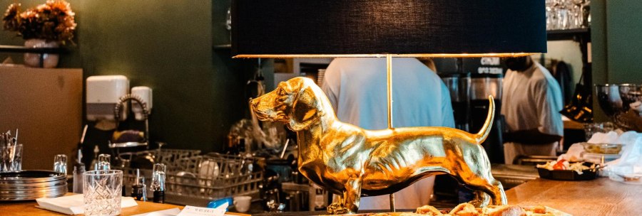 Eine goldene Dackel-Lampe steht auf einer Theke in einem Caf&eacute;. Im Hintergrund sind Gl&auml;ser und eine Person zu sehen., &copy; Stuttgart Marketing GmbH - Sarah Schmid