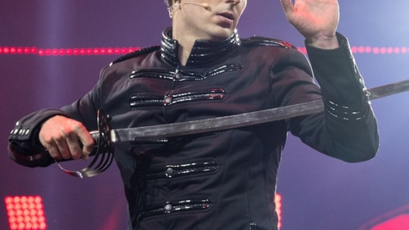 A man in a black jacket performs a sword illusion on a stage. He holds a sword and raises his hand as if he is performing magic., &copy; Renitenztheater Stuttgart e.V.