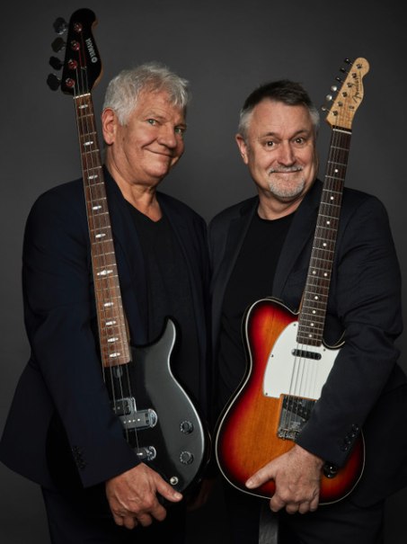 Two men in dark clothing hold electric guitars in front of a gray background and smile into the camera., © Renitenztheater Stuttgart e.V. Two men in dark clothing hold electric guitars in front of a gray background and smile into the camera., © Renitenztheater Stuttgart e.V.
