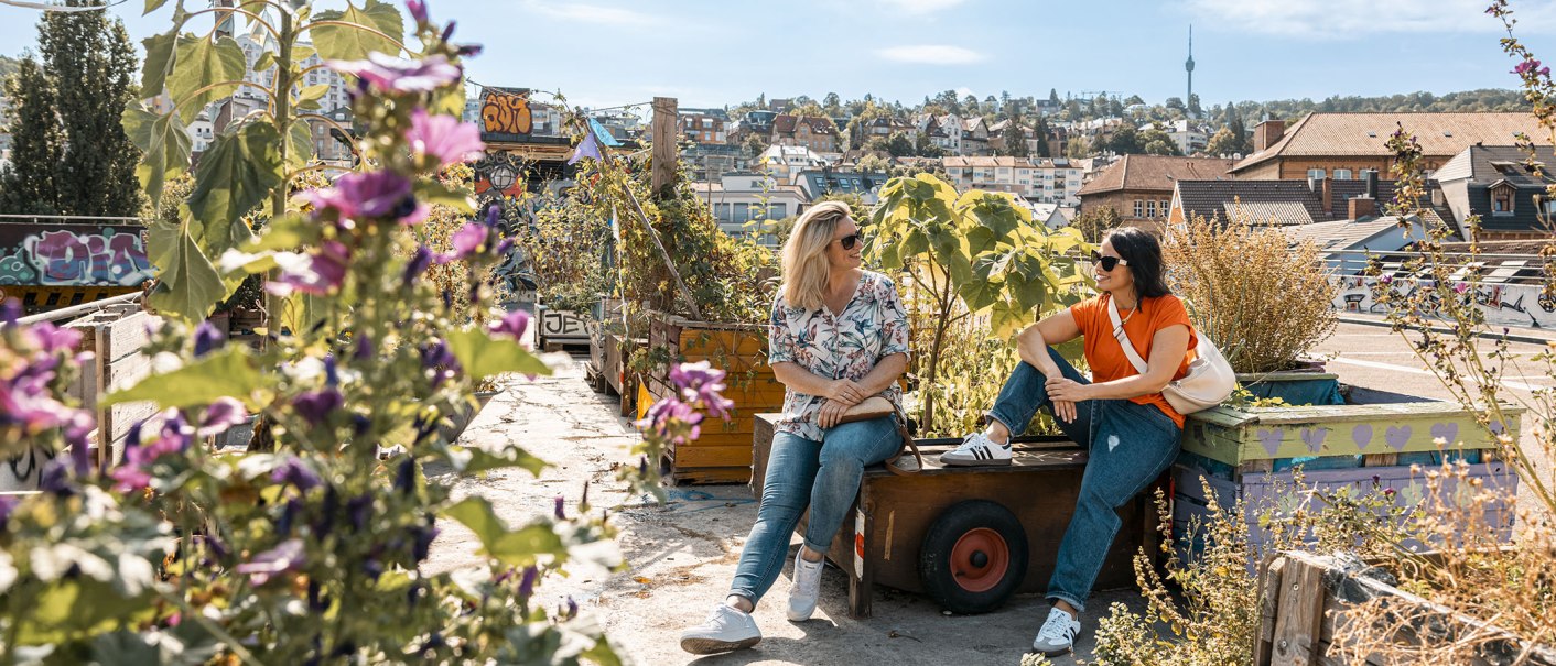 Zwei Frauen sitzen in einem urbanen Dachgarten mit Pflanzen und genießen den Ausblick auf die Stadt im Hintergrund., © © Stuttgart-Marketing GmbH, Sarah Schmid Zwei Frauen sitzen in einem urbanen Dachgarten mit Pflanzen und genießen den Ausblick auf die Stadt im Hintergrund., © © Stuttgart-Marketing GmbH, Sarah Schmid