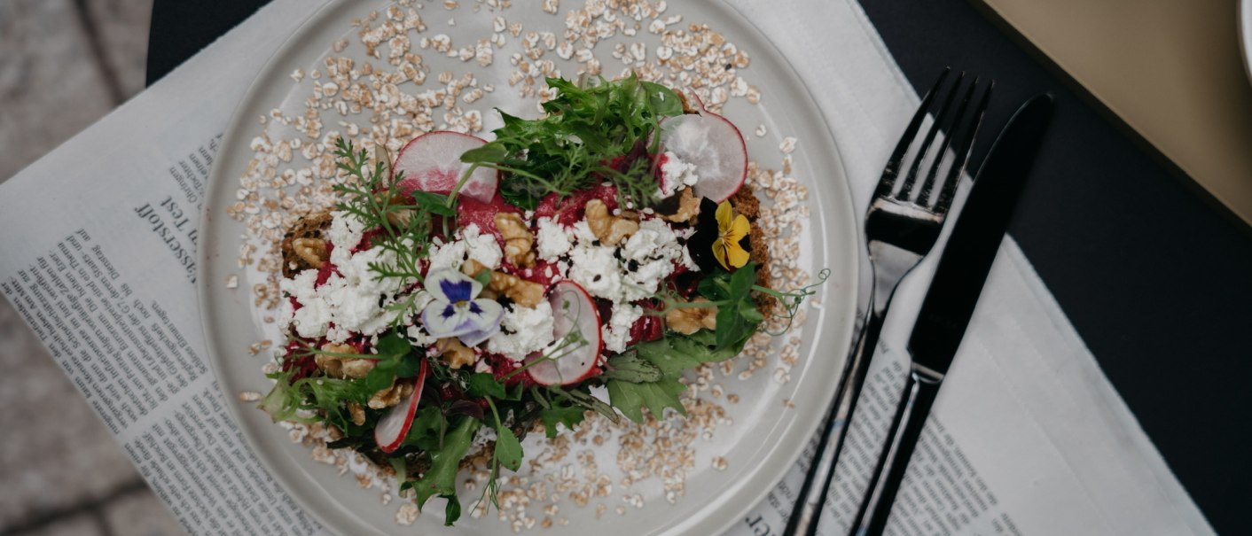 Ein dekorativer Salat mit Radieschen, Nüssen und essbaren Blüten auf einem Teller, serviert auf einer Zeitung., © Fritz, Stuttgart Ein dekorativer Salat mit Radieschen, Nüssen und essbaren Blüten auf einem Teller, serviert auf einer Zeitung., © Fritz, Stuttgart