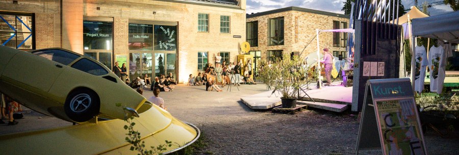 Evening event at the Kunstverein Wagenhalle with an art installation of a yellow car and people sitting in the square watching a performance., © Atelier Oradoro Evening event at the Kunstverein Wagenhalle with an art installation of a yellow car and people sitting in the square watching a performance., © Atelier Oradoro