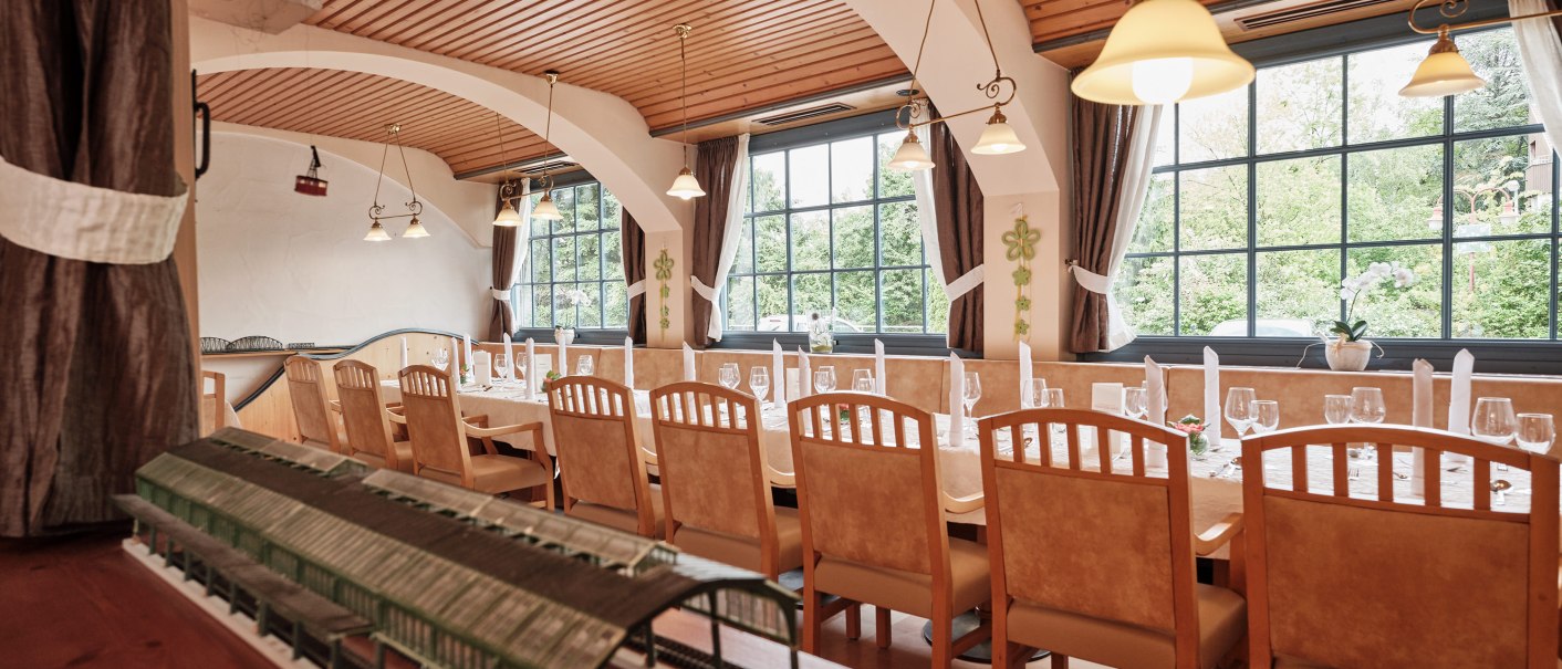 A restaurant with long, set tables, a wooden ceiling, and large windows. A model train can be seen in the foreground., © Badhotel Restaurant Stauferland A restaurant with long, set tables, a wooden ceiling, and large windows. A model train can be seen in the foreground., © Badhotel Restaurant Stauferland