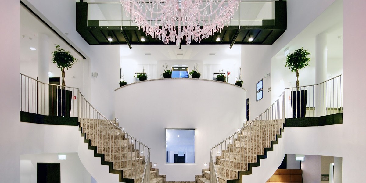 Elegante Lobby mit symmetrischer Treppe, großem Kronleuchter und Pflanzen. Moderne, helle Gestaltung mit Glas und Metallakzenten., © Arcotel Hotels