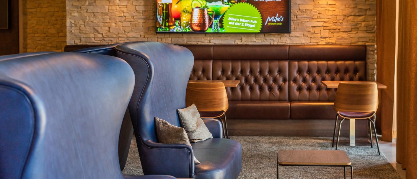 Moderne Hotelbar mit blauen Sesseln, braunen Ledersofas und einem Bildschirm mit Werbung für Mike's Urban Pub an der Wand., © Hilton Garden Inn Stuttgart Neckarpark