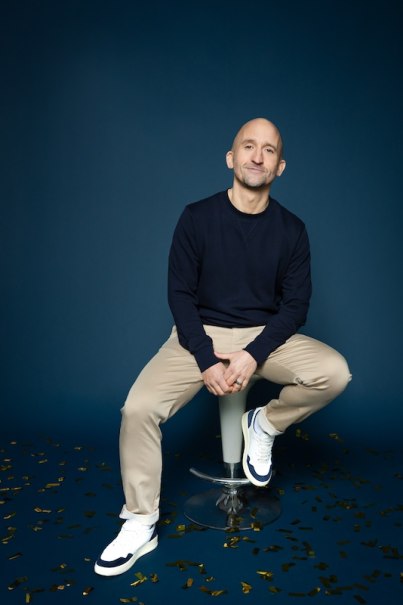 Person sitzt auf einem Hocker vor blauem Hintergrund, trägt dunklen Pullover und helle Hose. Goldene Konfetti liegen auf dem Boden., © Rosenau Kultur e.V. Person sitzt auf einem Hocker vor blauem Hintergrund, trägt dunklen Pullover und helle Hose. Goldene Konfetti liegen auf dem Boden., © Rosenau Kultur e.V.