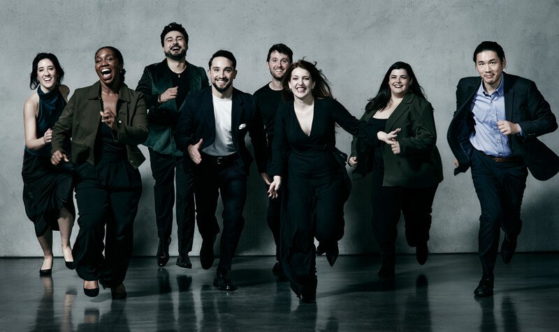 Acht Personen in eleganter Kleidung laufen lächelnd auf die Kamera zu. Der Hintergrund ist eine graue Wand, der Boden reflektiert leicht., © Württembergische Staatstheater Stuttgart Acht Personen in eleganter Kleidung laufen lächelnd auf die Kamera zu. Der Hintergrund ist eine graue Wand, der Boden reflektiert leicht., © Württembergische Staatstheater Stuttgart