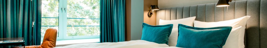 Modern hotel room with large bed, blue pillows and curtains. A window offers a view of the greenery. There is a desk and chair next to it., &copy; Motel One - Stuttgart Hauptbahnhof