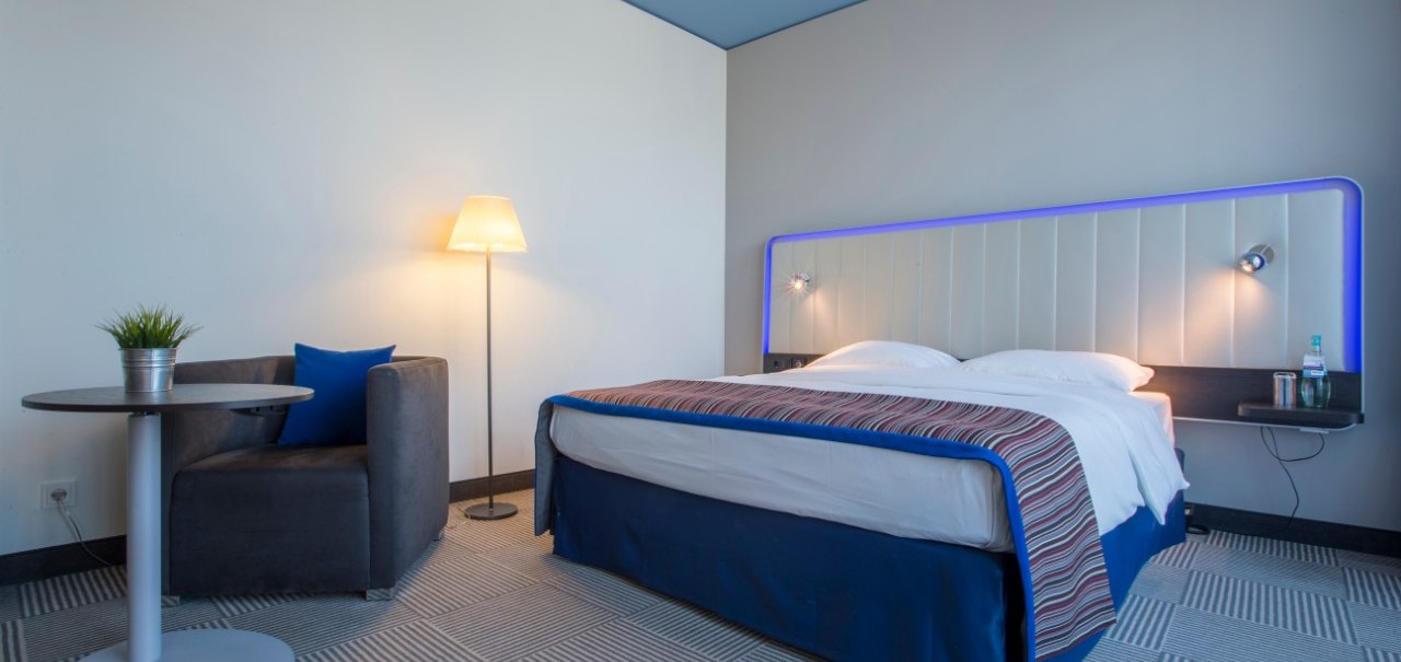 Modernes Gästezimmer mit Doppelbett, blauem Kopfteil, Sessel, kleinem Tisch mit Pflanze und Stehlampe auf gemustertem Teppichboden., © Park Inn by Radisson Stuttgart
