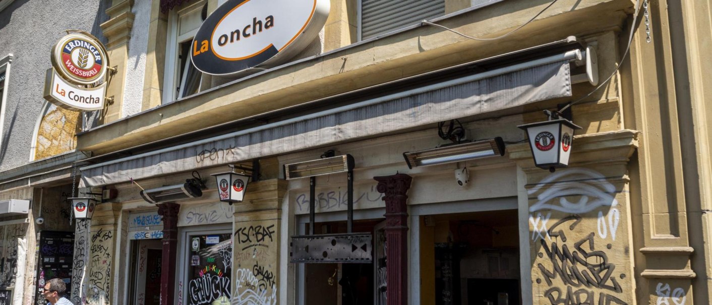 Fassade einer Bar namens La Concha mit Graffiti und Schildern von Erdinger Weißbräu. Die Außenwände sind mit Graffiti verziert., © SMG, Sarah Schmid Fassade einer Bar namens La Concha mit Graffiti und Schildern von Erdinger Weißbräu. Die Außenwände sind mit Graffiti verziert., © SMG, Sarah Schmid