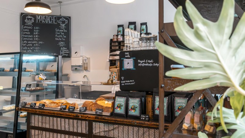 Ein gemütliches Café mit Theke, Gebäckauslage und Getränkekarte. Pflanzen schmücken den Raum, und eine Tafel zeigt das Menü an., © SMG, Sarah Schmid