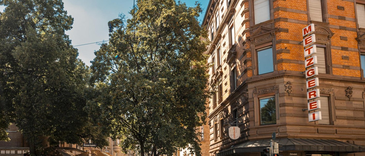 Eine Straßenecke mit einer Metzgerei und einem Café am Bismarckplatz. Menschen sitzen draußen unter Bäumen, Fahrräder sind abgestellt., © SMG, Sarah Schmid