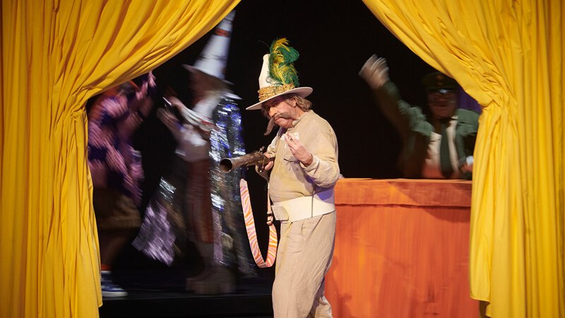 Ein Schauspieler in einem Kostüm mit Hut und Feder steht auf einer Bühne vor einem gelben Vorhang. Im Hintergrund sind weitere Darsteller zu sehen., © Württembergische Staatstheater Stuttgart Ein Schauspieler in einem Kostüm mit Hut und Feder steht auf einer Bühne vor einem gelben Vorhang. Im Hintergrund sind weitere Darsteller zu sehen., © Württembergische Staatstheater Stuttgart