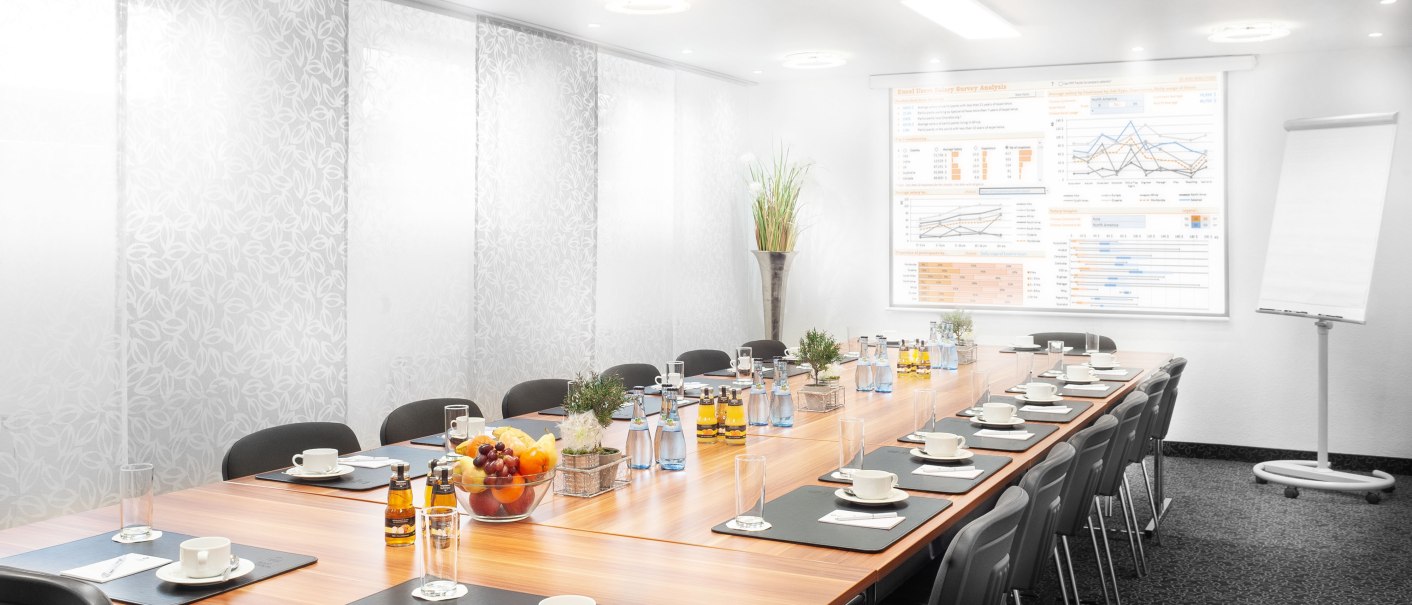 A modern conference room with a long table set with cups, glasses, and water bottles. A presentation on the wall, with a flip chart next to it., &copy; TOMAS
