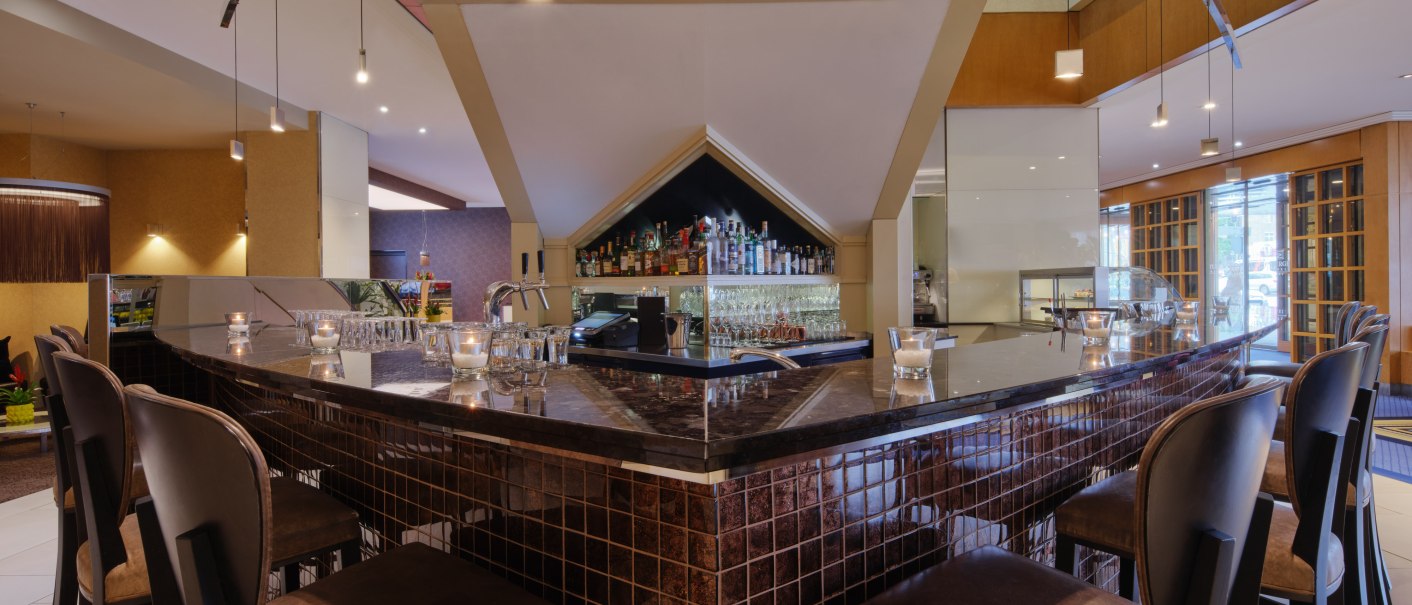 Modern bar with dark wooden furniture, stools and stylish lighting. Bottles and glasses are visible behind the bar., © Steigenberger Graf Zeppelin Modern bar with dark wooden furniture, stools and stylish lighting. Bottles and glasses are visible behind the bar., © Steigenberger Graf Zeppelin