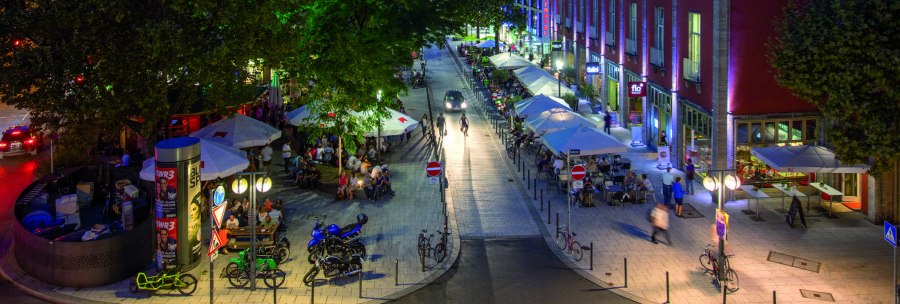 Lively street at night with cafés and people. Trees and colorful lights create a lively atmosphere. Bicycles and cars can be seen., © Stuttgart-Marketing GmbH