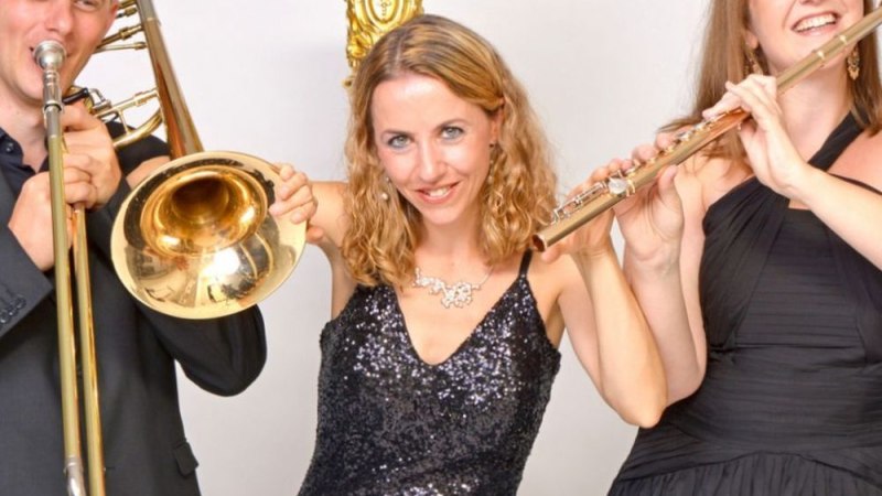 Drei Musiker in Abendkleidung posieren mit Posaune und Flöte vor einem goldenen Dekor. Sie lächeln und wirken fröhlich., © Trio Glissando Stuttgart