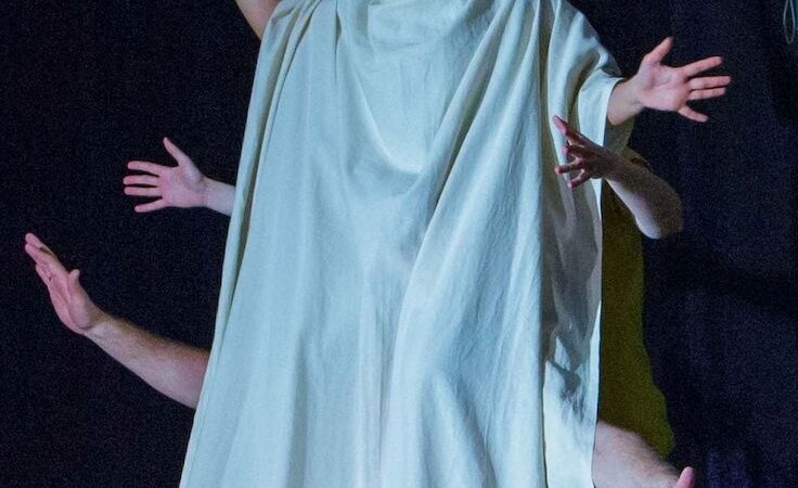 Eine Frau in einem wei&szlig;en Gewand mit mehreren H&auml;nden, die aus dem Gewand ragen, auf einer B&uuml;hne vor dunklem Hintergrund., &copy; Theater Atelier